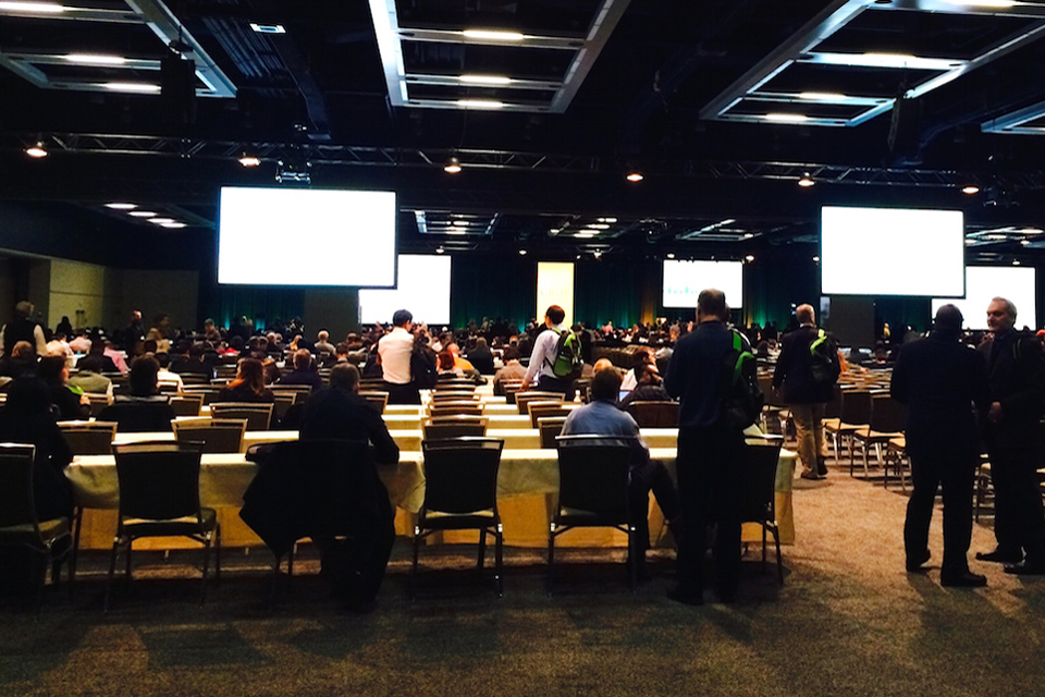 Many people sitting and standing at conference, with multiple screens hanging from ceiling. 