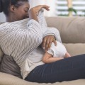 Woman sitting on couch looking under her shirt at infant underneath breastfeeding.