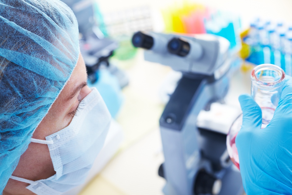 Lab technician in mask, near a microscope, looking at a flask of liquid. 