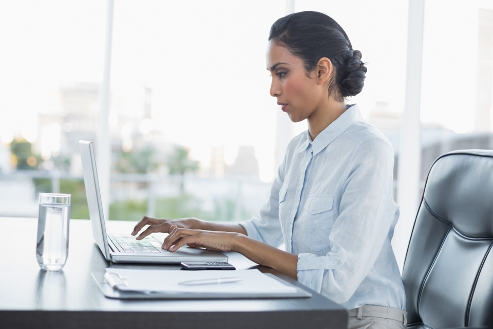 Profile of woman using laptop at desk with glass of water and phone on it near large window.