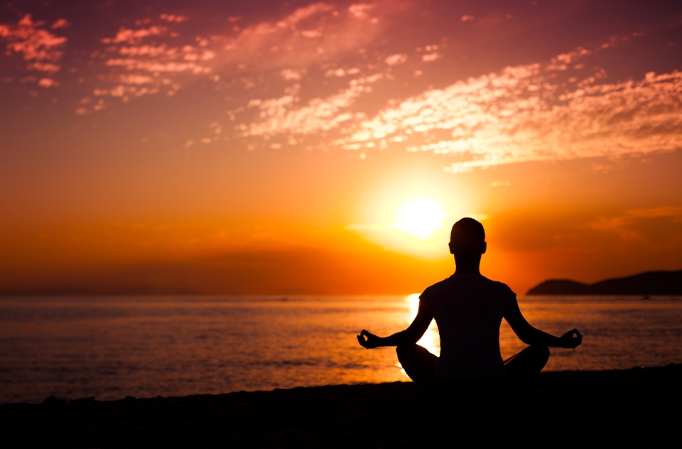 Silhouette of back of person in seated yoga pose near sea at sunset.