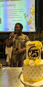 Rosaline M'bayo holding a microphone with a sign behind her and a cake in the foreground decorated with &quot;25&quot;.