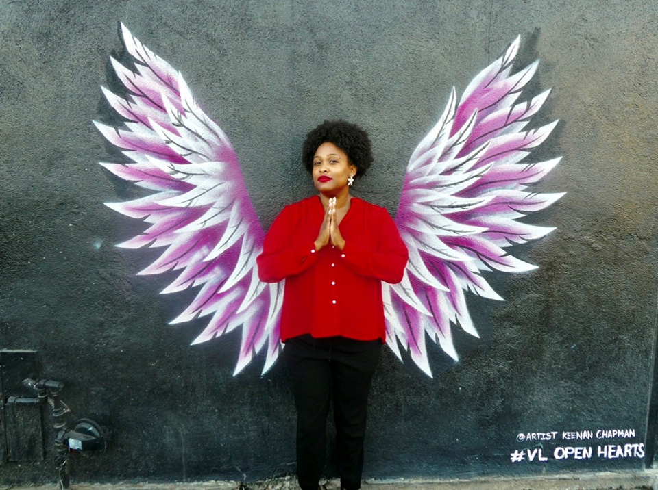 Porchia Dees, standing with palms together, in front of painting of wings on wall by Keenan Chapman.