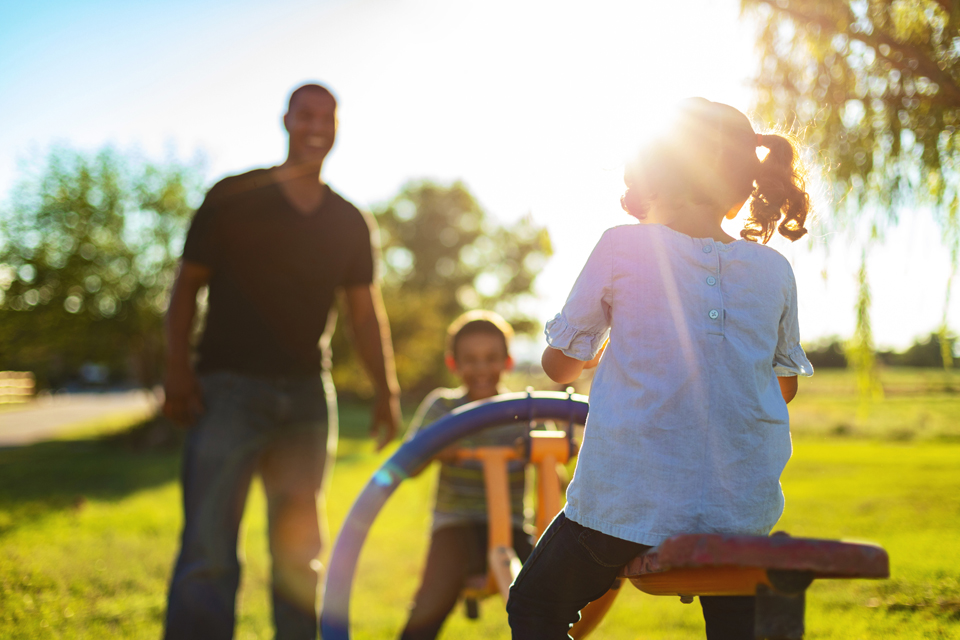 Hombre sonriendo en el parque con dos niños en un balancín.