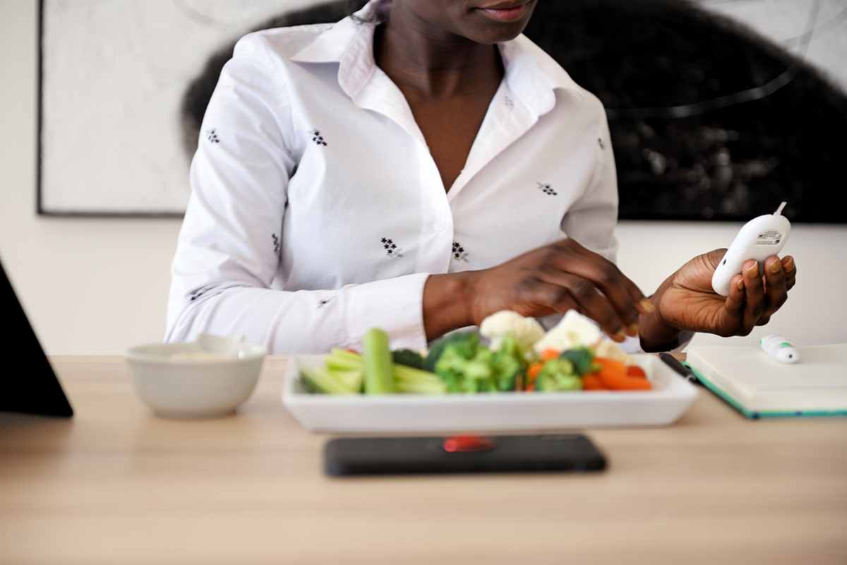 Woman sitting at table with a plate of vegetables and a bowl in front of her and holding and looking at blood sugar monitor.