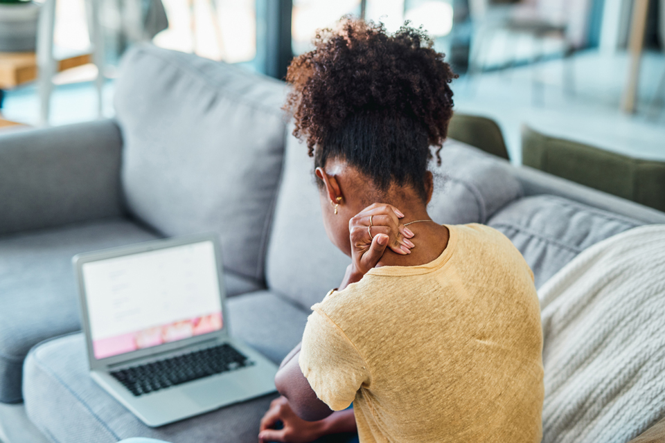 Back of woman holding the back of her neck and looking at a laptop.