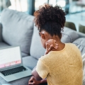 Back of woman holding the back of her neck and looking at a laptop.