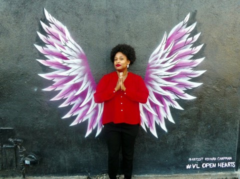 Porchia Dees, standing with palms together, in front of painting of wings on wall by Keenan Chapman.