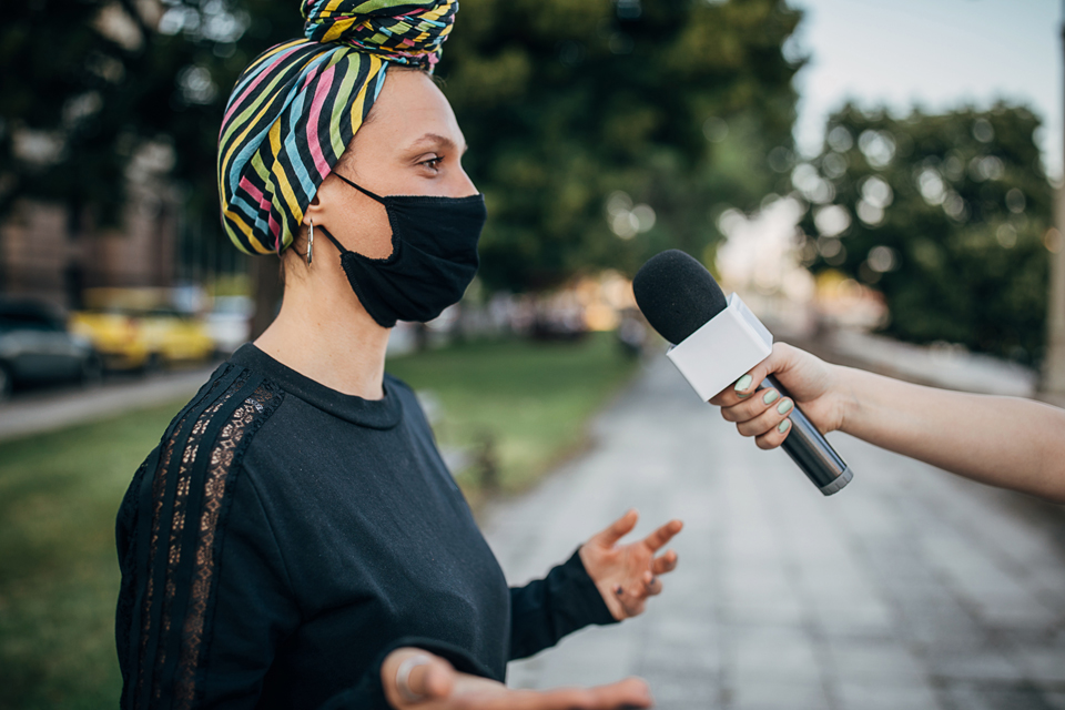 Profile of person in headwrap and mask with a microphone being held to her face by someone's hand..